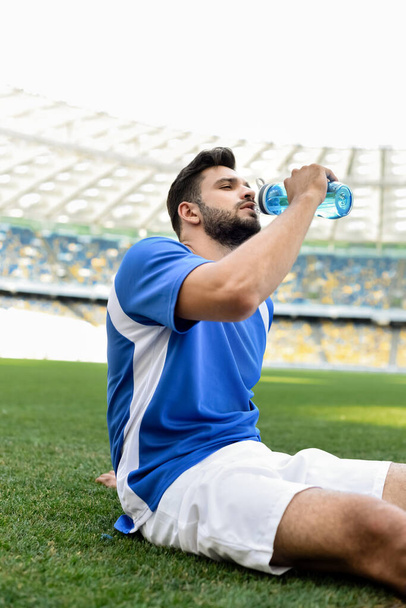 professionele voetballer in blauw en wit uniform zittend op voetbalveld en drinkwater in het stadion - Foto, afbeelding