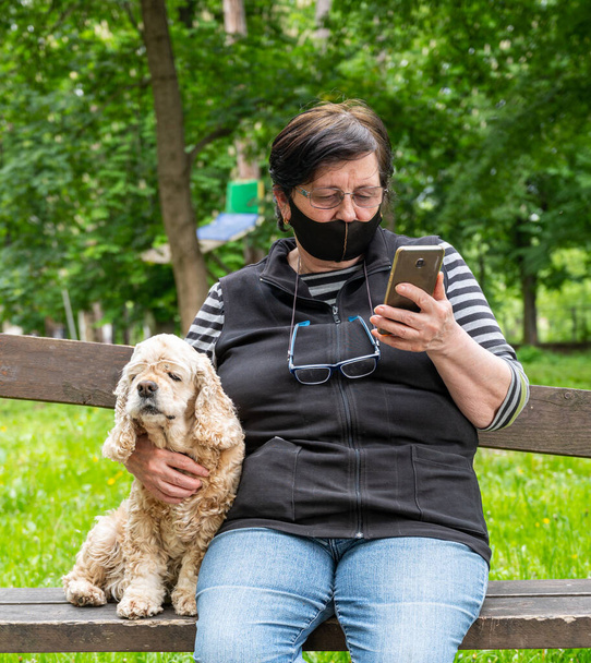 Donna anziana in maschera protettiva seduta su una panchina con un cane nel parco
 - Foto, immagini