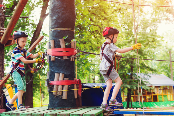 A gyerekek a fák között haladnak el a kötélpálya mellett. Fiatal testvérek másznak magas kötélpályán a Adventure Parkban. Gyerekek az erdő kalandparkjában. Sisakos gyerekek, extrém sport. Nyári tábor. - Fotó, kép