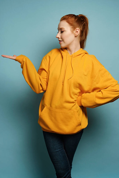 The red-haired girl stands on a blue background in a yellow hoodie, pointing her palm to the side and turning her head profile - Fotoğraf, Görsel