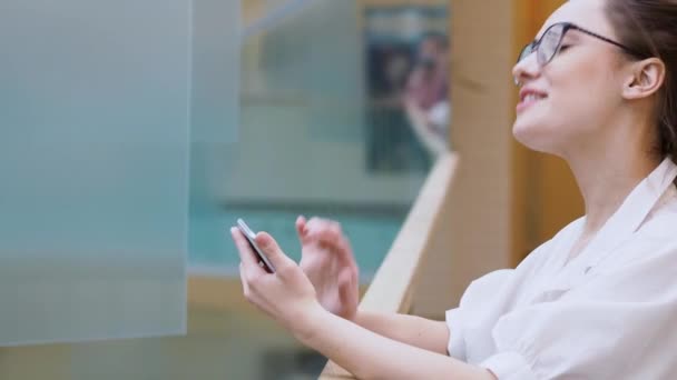 young business lady uses a smartphone. surprised girl in white shirt and glasses makes a phone call. - Materiał filmowy, wideo
