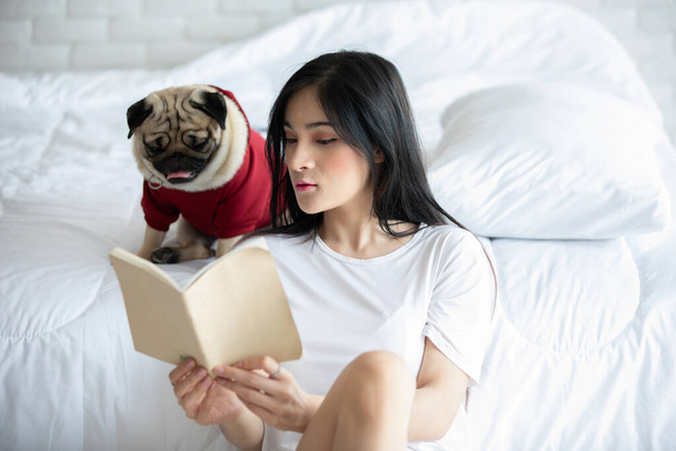 Attraktive asiatische junge Frau liest Buch, während ihr Hund Mops im gemütlichen Schlafzimmer brütet, entspannt und bequem im Urlaub, Erholung mit Hund - Foto, Bild