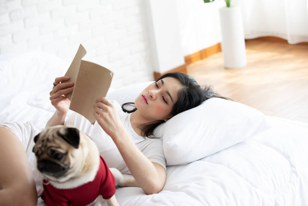 Приваблива азіатська молода жінка, яка читає книжку, а її собака виводить породу в затишній спальні, відпочиває і комфортно на канікулах, відпочиває з собачою концепцією - Фото, зображення