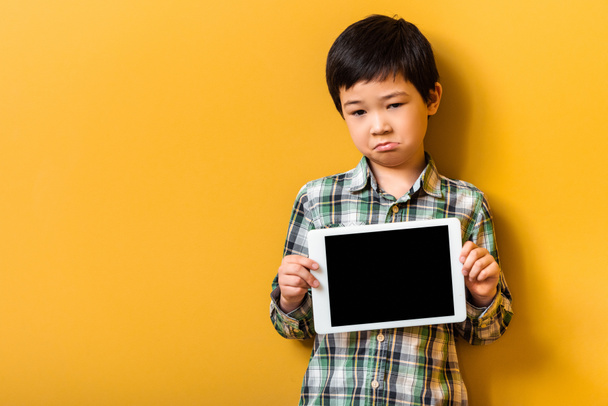 upset asian boy holding digital tablet with blank screen on yellow - Photo, image