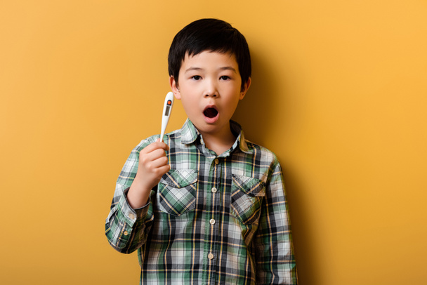 surprised asian boy holding thermometer on yellow - Φωτογραφία, εικόνα