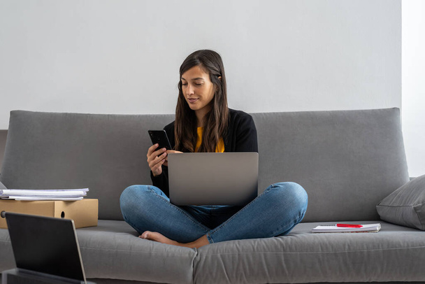 Giovane donna con computer portatile e telefono telelavoro sul divano a casa sua
 - Foto, immagini