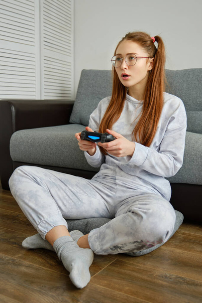 Redhead girl in pajamas sits on the floor with a game joystick and looks in surprise in front of her - Foto, imagen