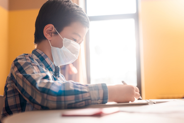 asiatischer Junge in medizinischer Maske schreibt und studiert zu Hause mit Sonnenlicht während der Quarantäne - Foto, Bild