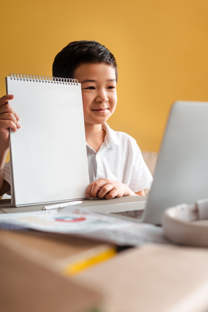 χαρούμενο ασιατικό αγόρι που σπουδάζει online με σημειωματάριο και φορητό υπολογιστή στο σπίτι κατά τη διάρκεια της καραντίνας - Φωτογραφία, εικόνα