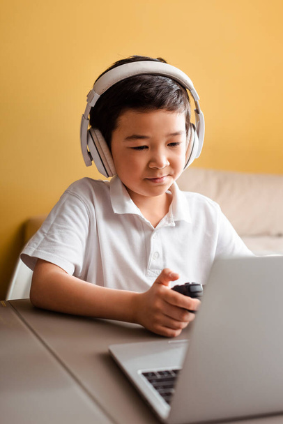 KYIV, UKRAINE - APRIL 22, 2020: asian boy playing video game with joystick and laptop on self isolation - Fotó, kép