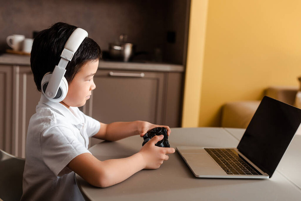 KYIV, UKRAINE - APRIL 22, 2020: asian boy playing video game with joystick and laptop with blank screen on quarantine - 写真・画像