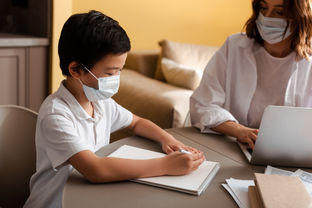 chico asiático lindo en máscara médica estudiando con la madre en casa en cuarentena. - Foto, imagen