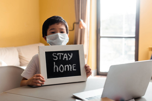 asiatischer Junge in medizinischer Maske mit Bleibe-zu-Hause-Schriftzug vor Laptop während Selbstisolation  - Foto, Bild