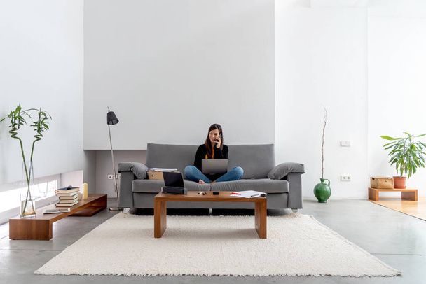 Young woman with laptop and phone telecommuting on the sofa at her home - Foto, immagini
