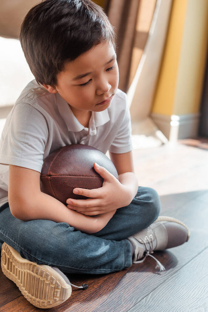 smutný asijský chlapec s rugby míč sedí na podlaze doma na vlastní izolaci - Fotografie, Obrázek