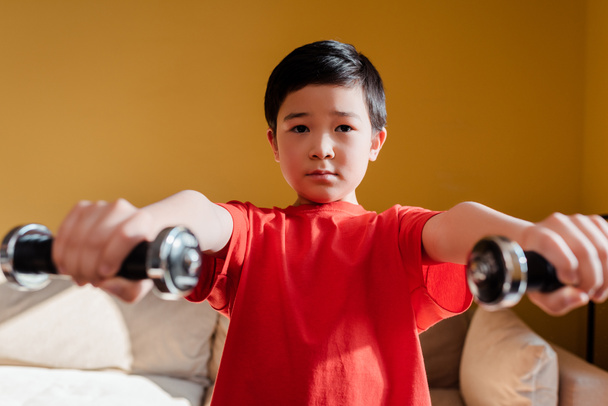 selektywne skupienie uwagi sportowego azjatyckiego chłopca ćwiczącego z hantlami w domu podczas kwarantanny - Zdjęcie, obraz
