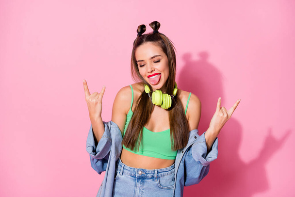 Photo of millennial lady showing finger horns sticking tongue out mouth listen earphones wear denim jacket green crop top naked belly isolated pastel pink color background - Foto, Imagem