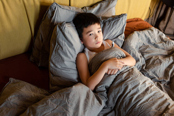 печально больной мальчик с термометром лежит в постели на самоизоляции - Фото, изображение