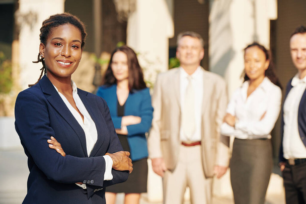 Beautiful confident successful businesswoman standing with arms folded and smiling at camera, her colleagues standing in background - Photo, Image