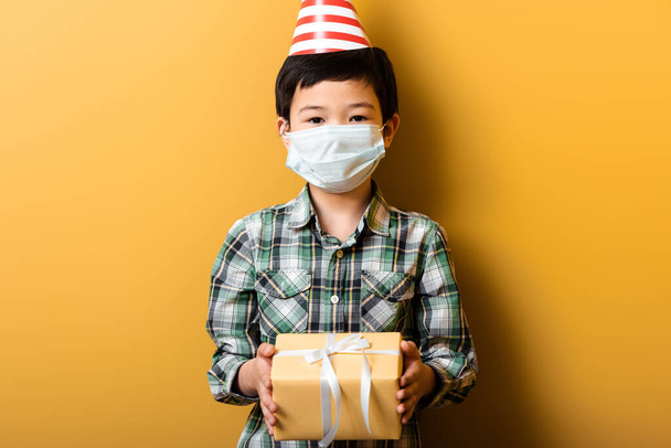 azjatycki chłopiec w stożek party i maska medyczna gospodarstwa urodziny prezent na żółty podczas samoizolacji - Zdjęcie, obraz