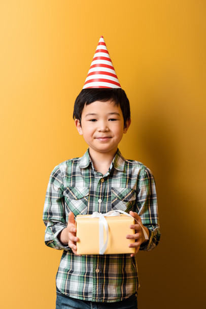 χαριτωμένο ασιατικό αγόρι στο κόμμα κώνο κρατώντας δώρο γενεθλίων στο κίτρινο  - Φωτογραφία, εικόνα