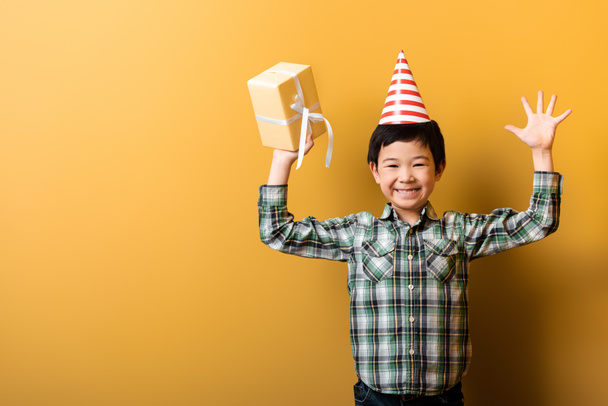 happy asian boy in birthday party cone holding present on yellow  - Foto, Imagen
