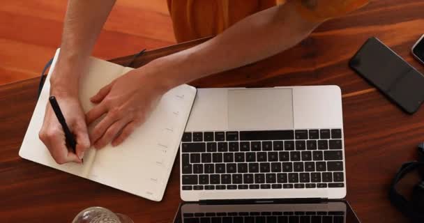 Hoge invalshoek van een blanke vrouw die tijd thuis doorbrengt tijdens het coronavirus, gesloten19, werkt aan een bureau, haar laptop gebruikt en aantekeningen maakt. Sociale afstand en zelfisolerend. - Video