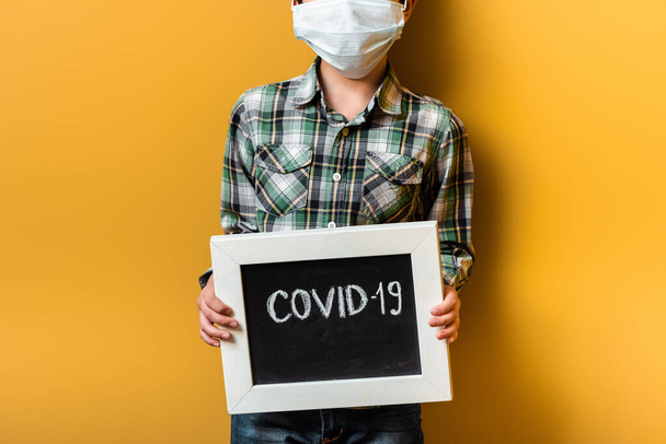 cropped view of boy in medical mask holding board with covid-19 sign on yellow during self isolation - Фото, изображение
