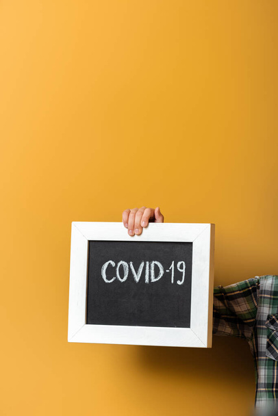 cropped view of kid holding board with covid-19 sign on yellow during quarantine - Foto, afbeelding