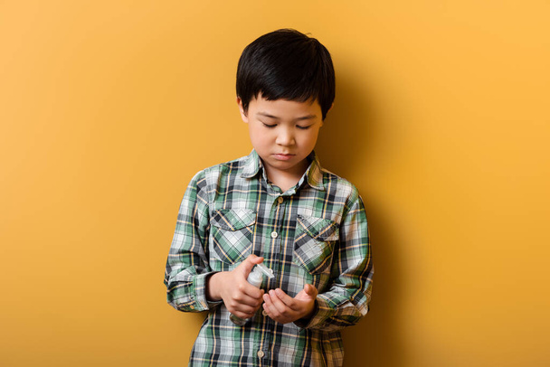 cute asian boy holding bottle with antiseptic on yellow during self isolation - Photo, Image