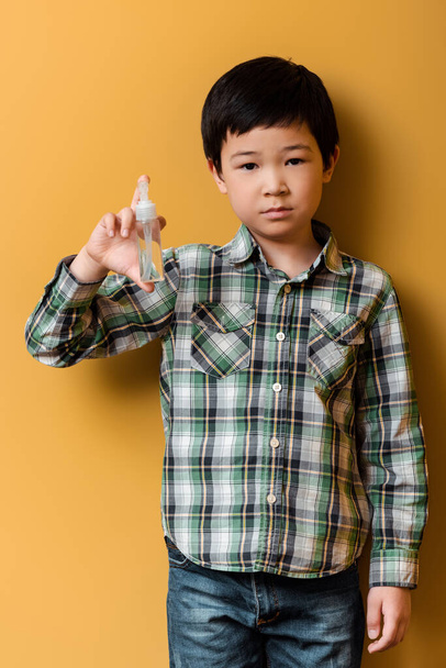 asian boy holding bottle with antiseptic on yellow during self isolation - Foto, Imagen