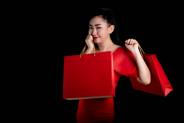 Portrait of a beautiful young Asia woman wearing a red dress holding credit card and shopping bags over a black background, sale concept, look at the camer - Foto, Imagem