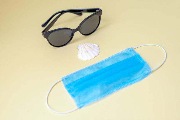 Sunglasses, medical face mask, seashell on a yellow background. Protection concept while traveling. - Photo, Image
