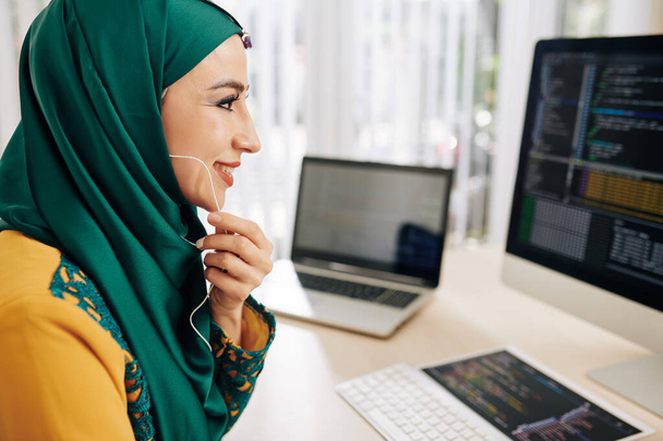 Iloinen hymyilevä muslimi nuori nainen, jolla on kuulokkeet, ohjelmointi tietokoneella ja puhuminen työtoverille
 - Valokuva, kuva
