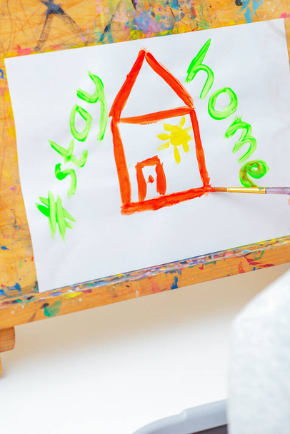 Hand van kind tekening rood huis en woorden Blijf thuis met aquarellen op de ezel thuis. Creatief concept voor kinderen. - Foto, afbeelding