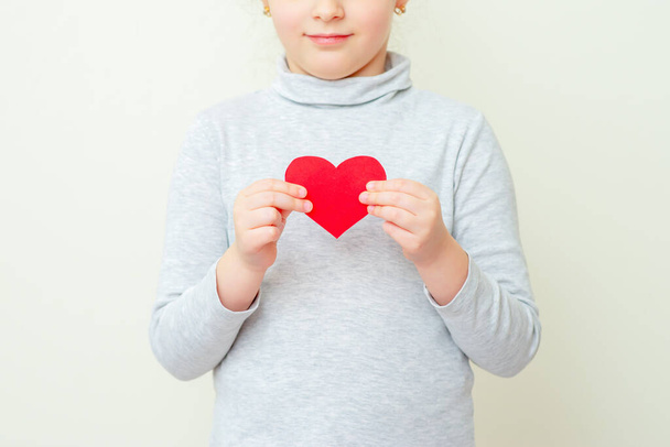 Little girl is holding red little paper heart standing on white background. Love concept. - Foto, Imagem