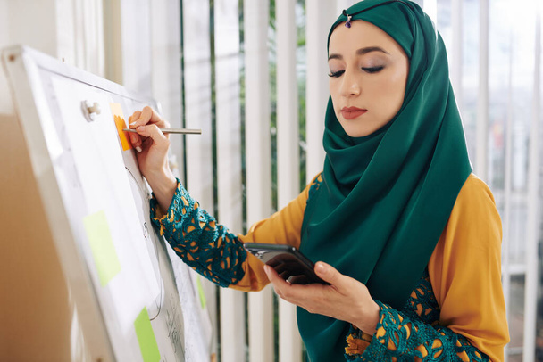 Geleneksel giysiler içinde genç, güzel, Müslüman bir iş kadını renkli çıkartmalar yazarken akıllı telefonu kontrol ediyor. - Fotoğraf, Görsel