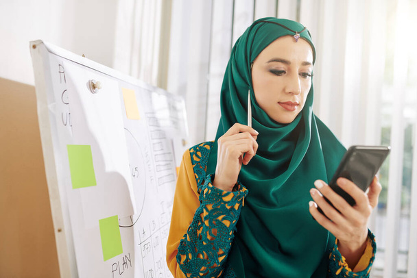Portret van jonge moslim zakenvrouw in traditionele kleding lezen artikel op smartphone scherm - Foto, afbeelding