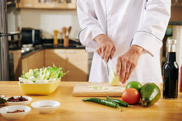 Zugeschnittenes Bild eines Mannes im Kochmantel, der beim Kochen zu Hause Zwiebeln schneidet - Foto, Bild