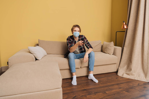 homem em máscara médica sentado no sofá e segurando controlador remoto
 - Foto, Imagem