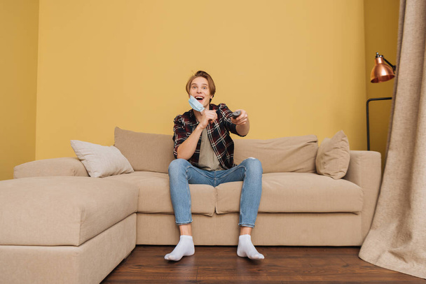 excited man with opened mouth touching medical mask, sitting on sofa and holding remote controller, end of quarantine concept  - Fotoğraf, Görsel