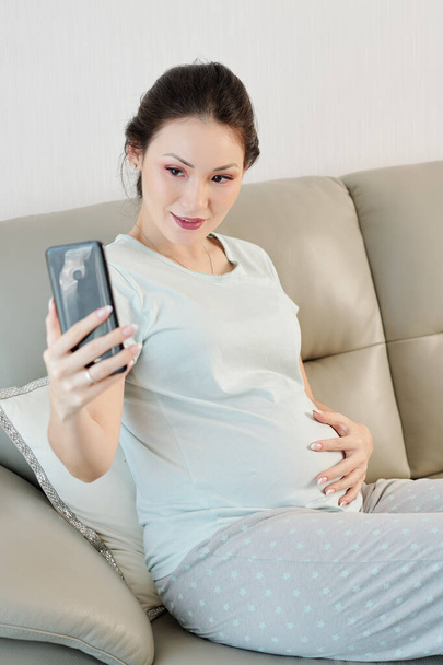 Beautiful young Vietnamese pregnant woman sitting on sofa and taking selfie for social media - Foto, imagen