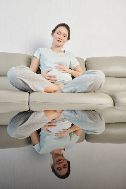Ευτυχισμένη νεαρή έγκυος γυναίκα κλείνει τα μάτια και αγγίζει την κοιλιά της για να αισθανθεί τις κινήσεις του μωρού - Φωτογραφία, εικόνα