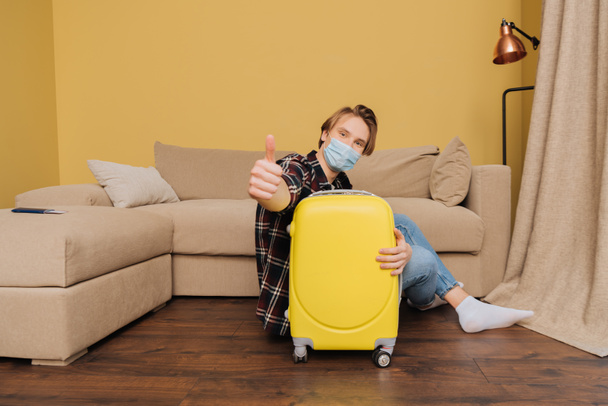 selective focus of man in medical mask sitting on floor near baggage and showing thumb up, end of quarantine concept - Φωτογραφία, εικόνα
