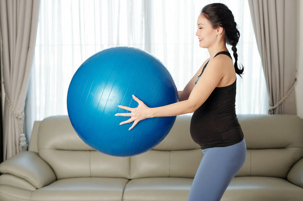 Jovem mulher grávida asiática feliz exercitando com bola de fitness em casa
 - Foto, Imagem