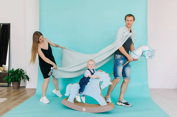 A handsome prince and a chic princess with a little son, a prince on a toy horse and a plaid raincoat are isolated on a blue background. Games at home in the interior paper background. - Fotoğraf, Görsel