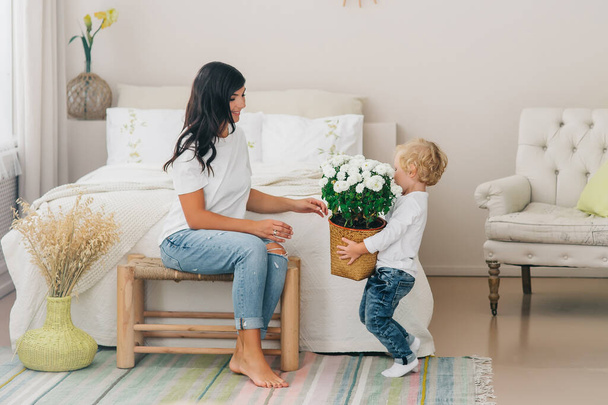 Blond cute boy gives mom a pot of white flowers. Beautiful cozy bedroom interior - Φωτογραφία, εικόνα
