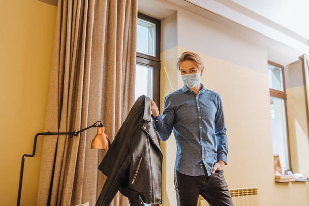 young man in medical mask holding jacket and standing with hand in pocket at home, end of quarantine concept  - 写真・画像