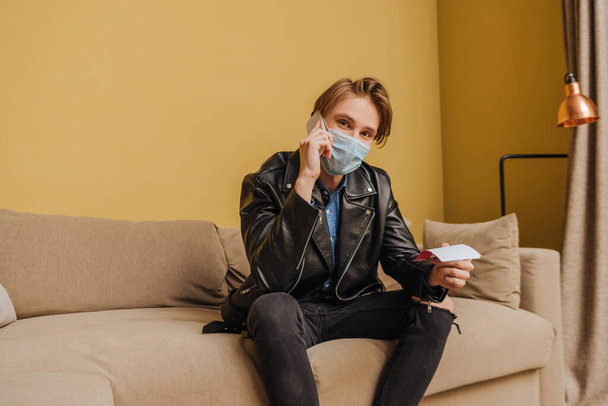 stylish man in medical mask and jacket holding tickets and talking on smartphone, end of quarantine concept  - Photo, Image