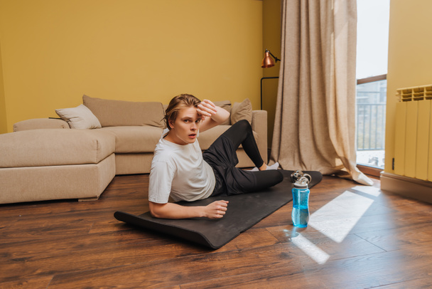 vermoeide man liggend op fitnessmat bij sportfles met water, einde quarantaine concept - Foto, afbeelding